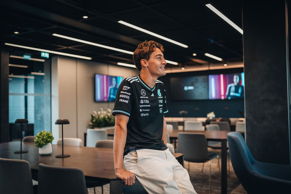 George Russell, piloto da equipe Mercedes-AMG PETRONAS Formula One Team, está sentado de forma descontraída sobre uma mesa em uma área de hospitalidade ou escritório moderna e elegante da equipe. Ele sorri e veste a camisa oficial do time com os logotipos dos patrocinadores visíveis, como Einhell, IWC e CrowdStrike. Telas ao fundo exibem sua imagem.