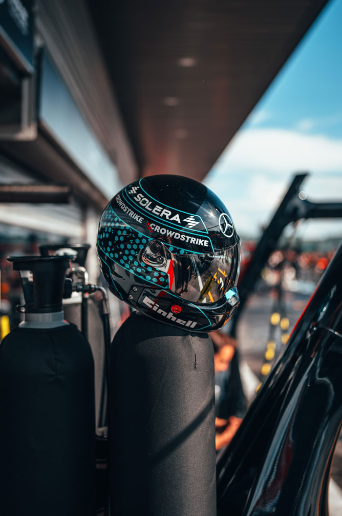 Um capacete da equipe Mercedes-AMG PETRONAS Formula One Team, com logotipos de patrocinadores bem visíveis, incluindo Einhell, CrowdStrike e Solera, está colocado sobre um cilindro de gás na pista dos boxes. O reflexo na viseira mostra o ambiente da garagem.