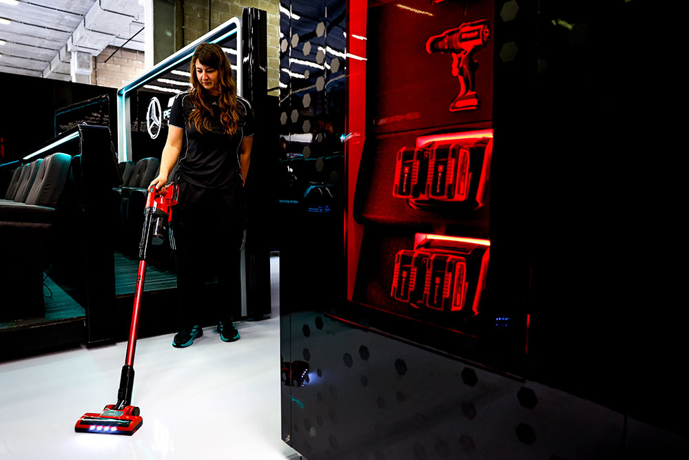 Uma mulher da equipe Mercedes-AMG PETRONAS Formula One Team limpa um piso branco brilhante com um aspirador de pó sem fio da Einhell. À direita, prateleiras iluminadas em vermelho exibem várias baterias Einhell Power X-Change e uma furadeira sem fio.