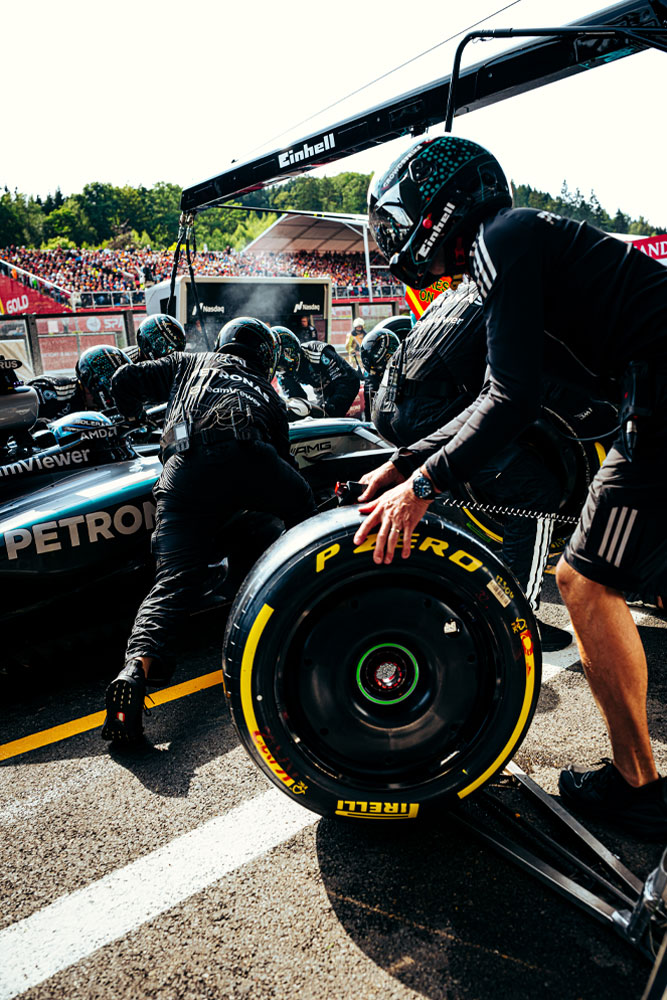 Mecânicos da equipe Mercedes-AMG PETRONAS Formula One Team realizam um pit stop. Um membro da equipe prepara um pneu Pirelli enquanto outros trabalham no carro. A marca Einhell é visível nos equipamentos acima do carro.