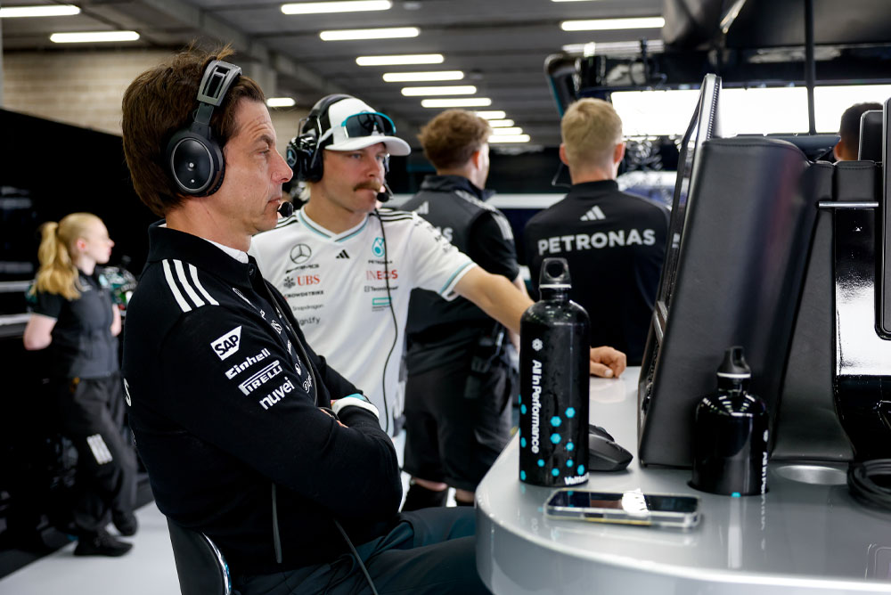 Toto Wolff e Valtteri Bottas, da equipe Mercedes-AMG PETRONAS Formula One Team, estão na estação de trabalho da equipe na garagem. Eles estão focados na estratégia de corrida, observando os monitores e se comunicando por meio de fones de ouvido.
