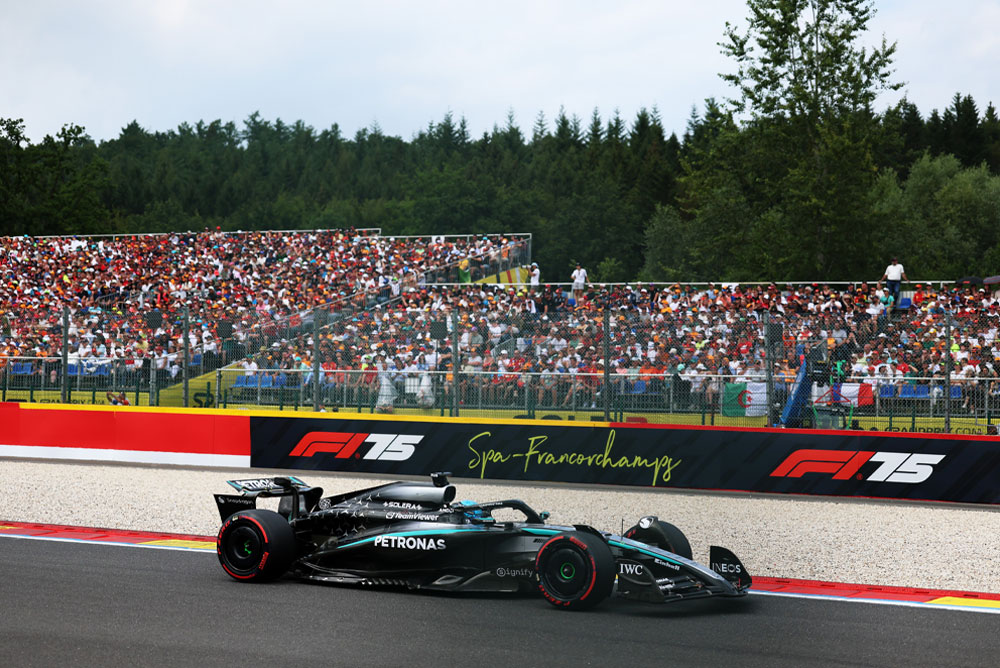 Um carro da equipe Mercedes-AMG PETRONAS Formula One corre no circuito de Spa-Francorchamps. Ao fundo, as arquibancadas estão lotadas de fãs acenando bandeiras internacionais. O logotipo de Spa-Francorchamps é exibido na barreira.