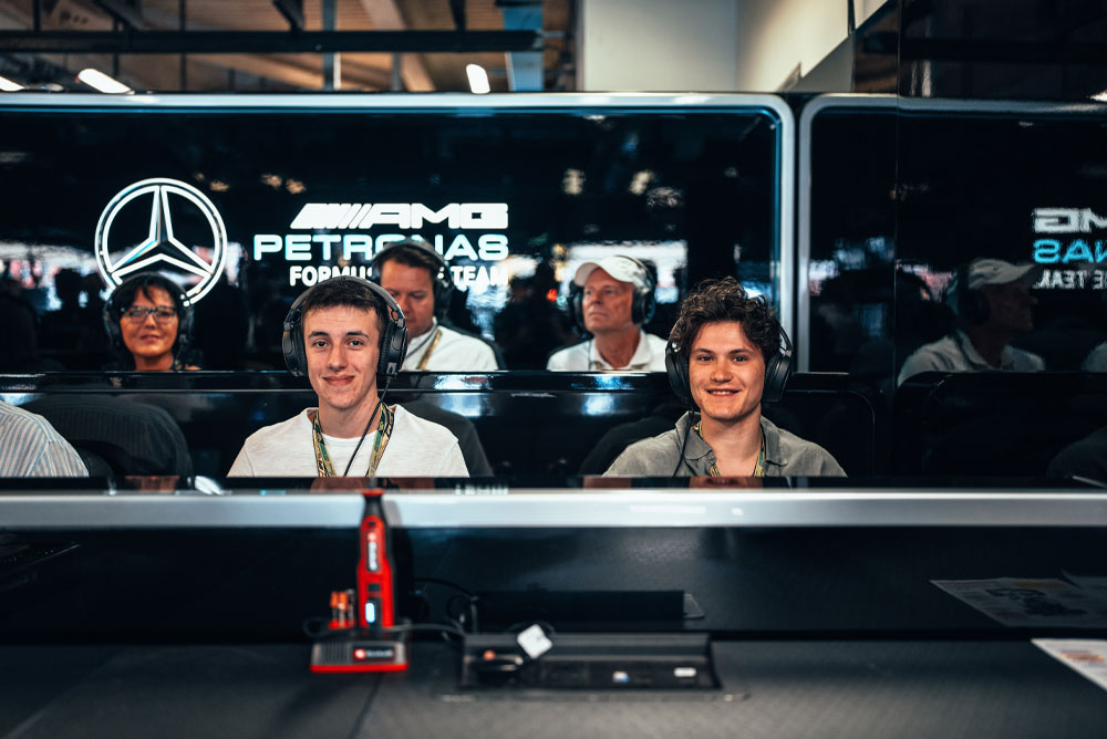 Dois homens estão sentados na garagem da equipe Mercedes-AMG PETRONAS Formula One, usando fones de ouvido e aproveitando a experiência nos bastidores. Um dispositivo vermelho da Einhell é visível em primeiro plano.