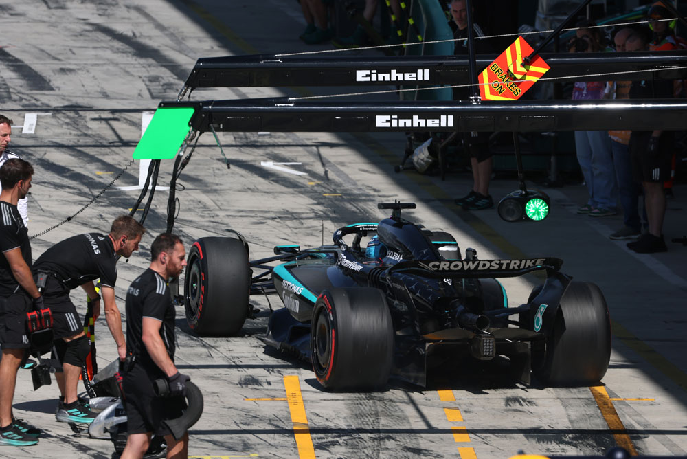 Um carro da equipe Mercedes-AMG PETRONAS Formula One está na pista dos boxes durante um pit stop. Vários membros da equipe estão ao redor do carro. A marca Einhell é visível acima da área dos boxes.