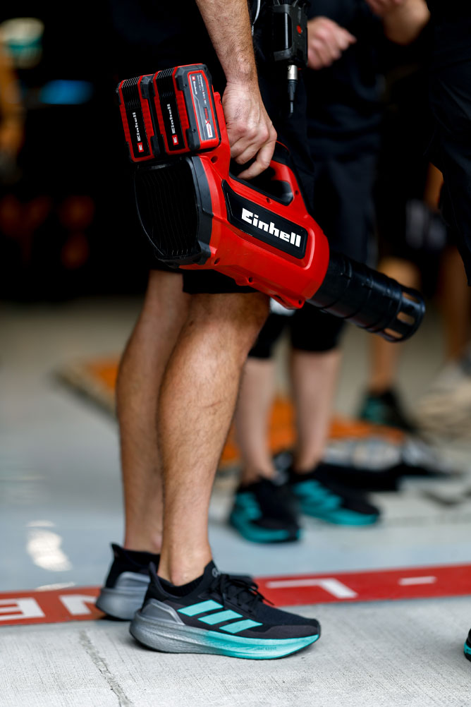 Um membro da equipe segura um soprador sem fio vermelho da Einhell com duas baterias Power X-Change. A cena acontece na área dos boxes durante um evento de Fórmula 1.