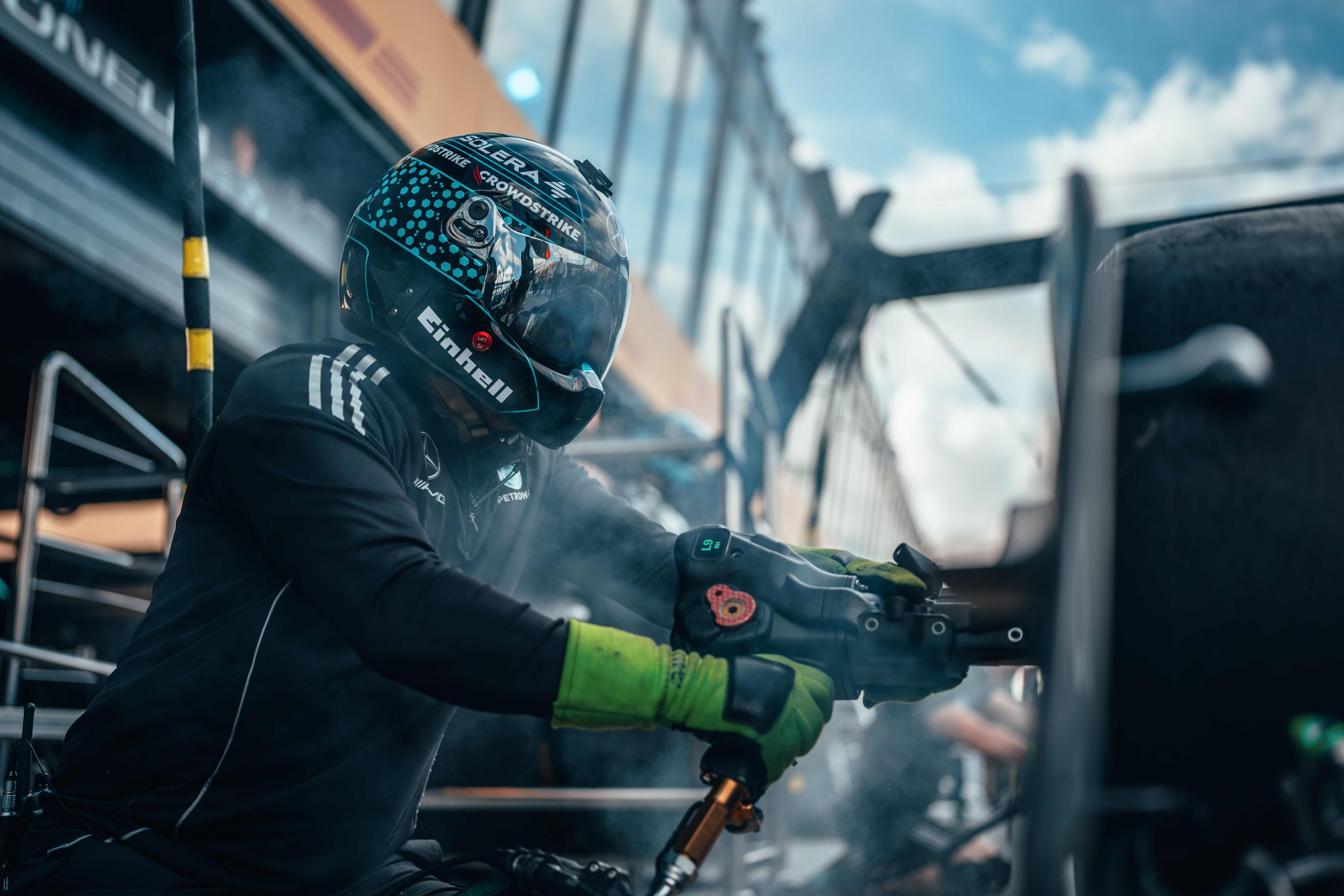 Um membro da equipe de boxes da Fórmula 1, usando capacete e luvas, utiliza uma pistola pneumática para rodas durante um pit stop, cercado por fumaça e equipamentos.