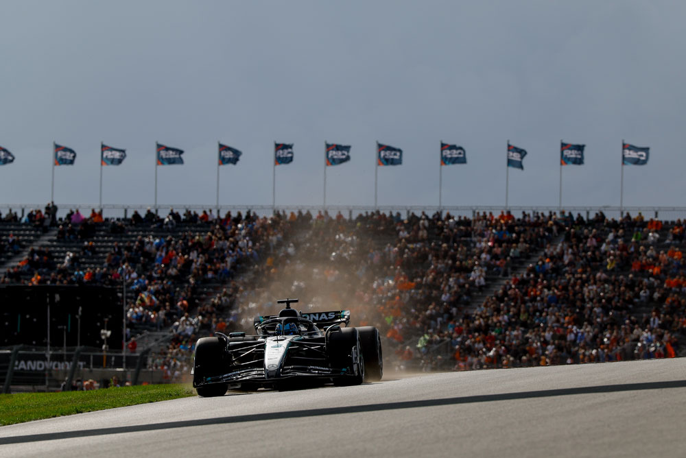 Um carro da Mercedes-AMG PETRONAS Formula One Team passa por arquibancadas lotadas durante uma corrida. Fãs acenam bandeiras ao fundo, criando uma atmosfera vibrante.