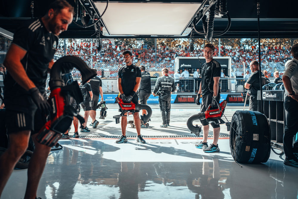 Mecânicos da equipe Mercedes-AMG PETRONAS Formula One se preparam na garagem com sopradores sem fio da Einhell antes de um pit stop. Ao fundo, as arquibancadas estão cheias de fãs.