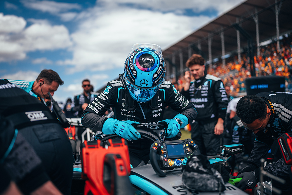 Piloto de Fórmula 1 sentado no carro rodeado pela equipe na grelha de largada.