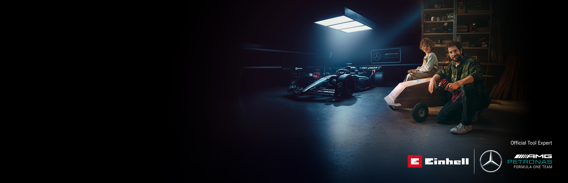 Um pai e uma criança numa oficina com um carro de rolamentos feito em casa. Ao fundo, um carro de Fórmula 1 da Mercedes-AMG PETRONAS e ferramentas Einhell.