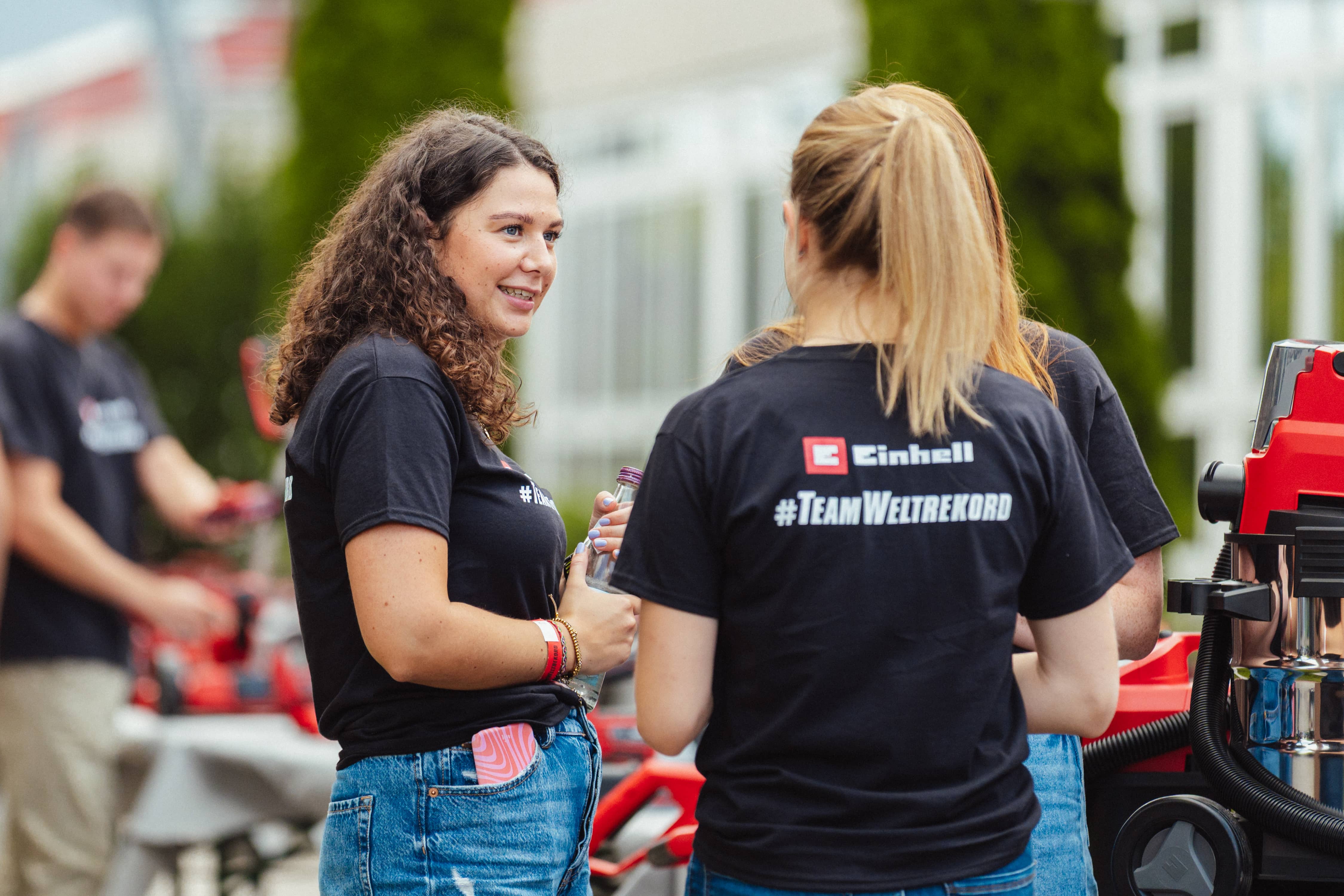 Duas jovens com t-shirts "#TeamWeltrekord" conversam em frente a ferramentas Einhell no exterior.