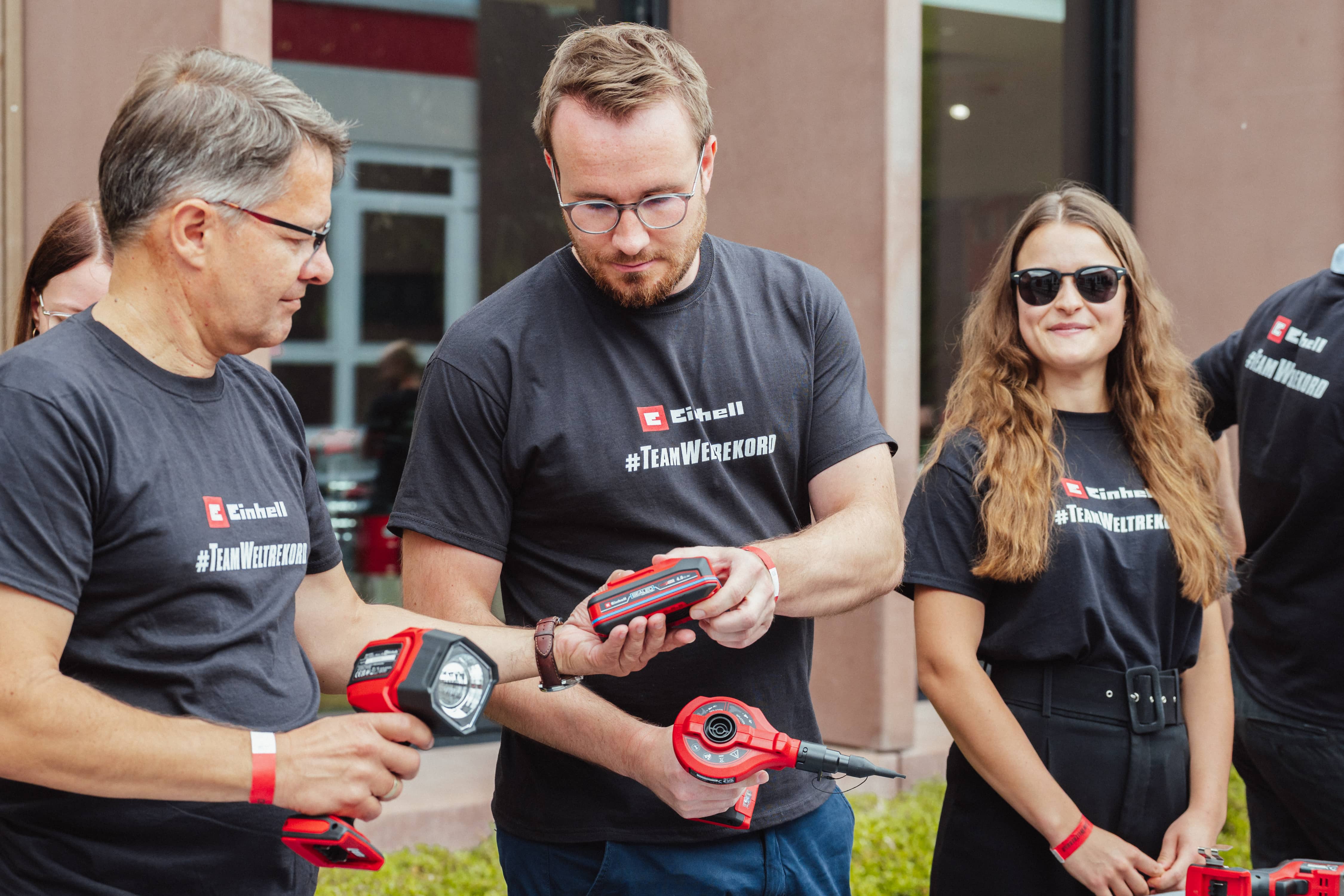 Três pessoas com t-shirts "#TeamWeltrekord" analisam em conjunto uma bateria Einhell SEALED.