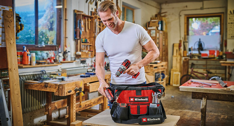 Uma bolsa de ferramentas, ao fundo um homem com uma chave de fenda sem fio numa oficina.